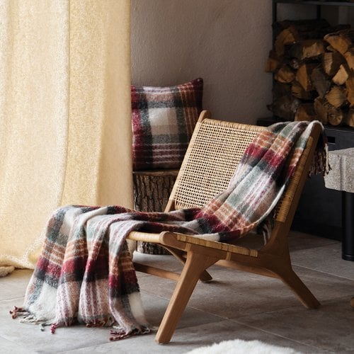 Fauteuil en bois et rotin avec un plaid écossais à motifs rouges et verts et un coussin assorti. Bûches de bois en arrière-plan.