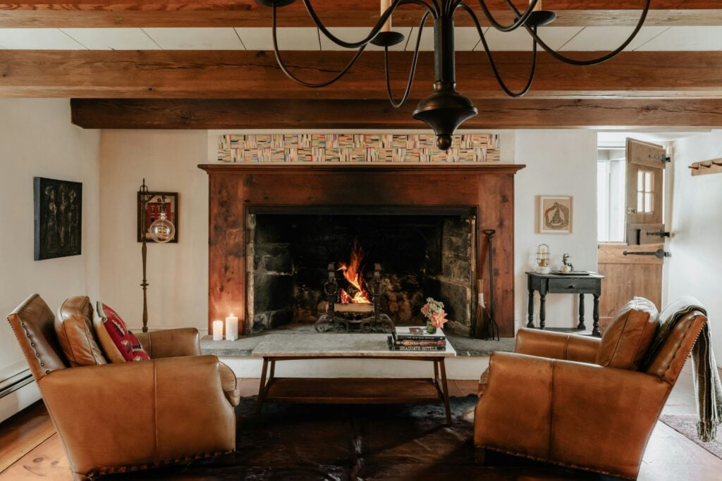 Salon rustique avec une grande cheminée en pierre et deux fauteuils club en cuir marron. Poutres en bois sombre au plafond et lustre en fer forgé.
