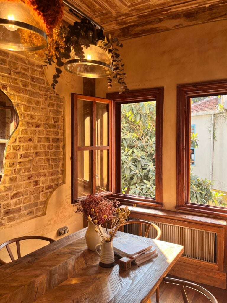 Coin repas dans une ambiance chaleureuse avec un mur de briques apparentes et des cadres de fenêtres en bois donnant sur la verdure. Table en bois verni.