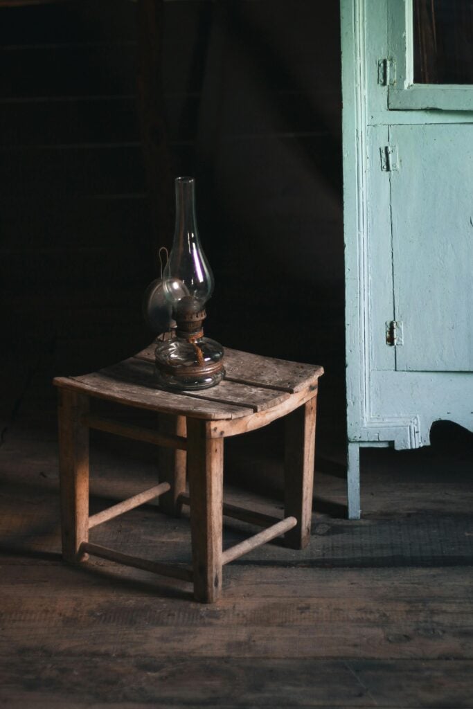 Lampe à huile vintage posée sur un petit tabouret rustique en bois dans une pièce sombre. Armoire en bois patiné bleu clair à droite.