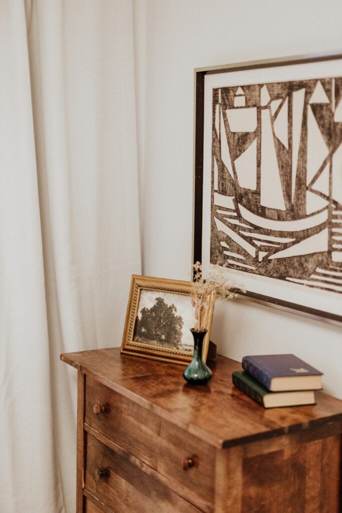 Commode en bois massif style modern farmhouse, avec diverses décorations poser sur le dessus, un rideau beige sur le fond et un tableau beige et marron au dessus de la commode.