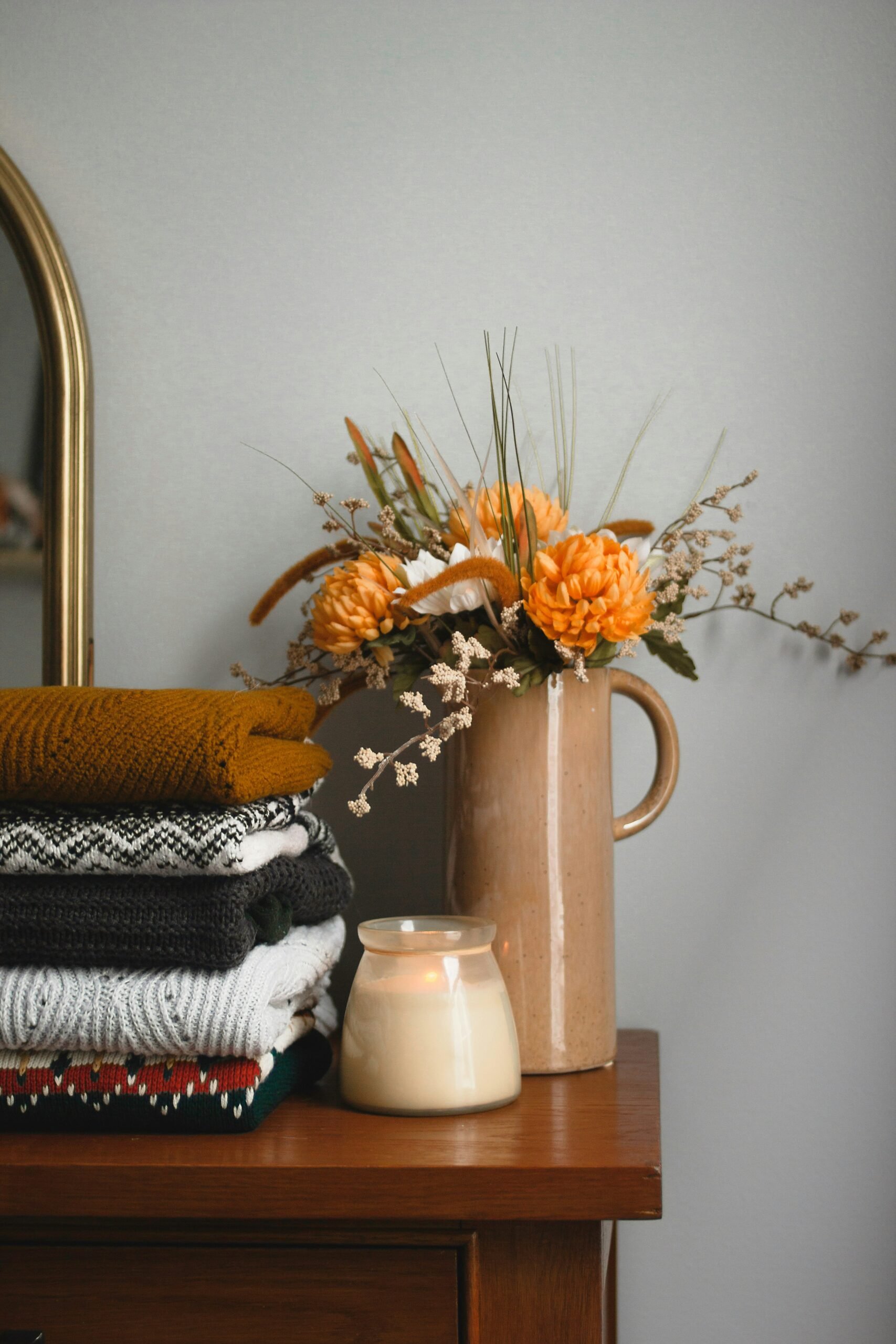 Mise en scène de décoration dans le style modern farmhouse, avec des pulls de couleurs différentes, une bougie et un pot en céramique avec un bouquet de fleurs au teinte automnale
